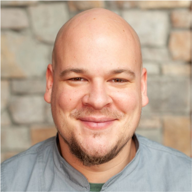 Against the stone wall backdrop, a person with a shaved head and goatee smiles warmly, perhaps savoring memories of dining at Cle Elum restaurants near Suncadia Resort.