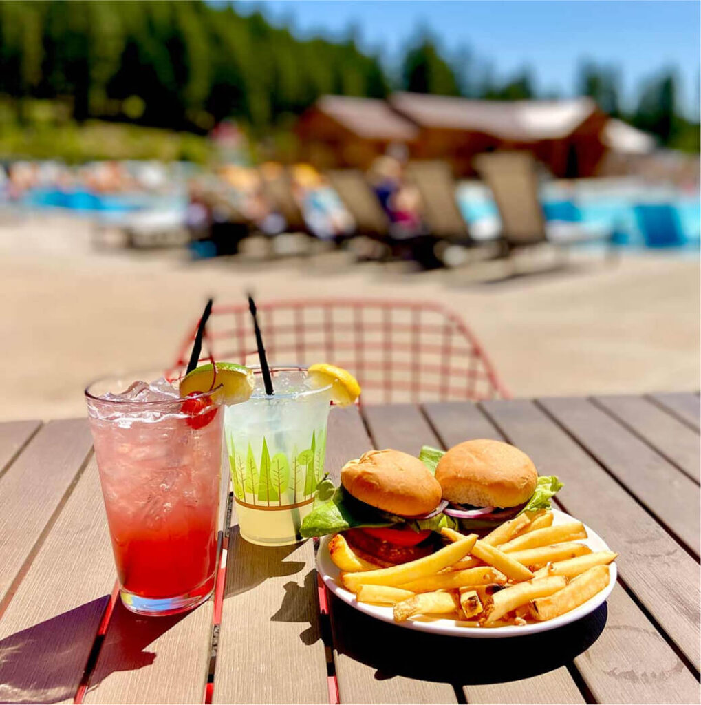 A plate with two burgers and fries, alongside two cocktails adorned with lemon and cherry garnishes, graces a table by the pool at Suncadia. Lounge chairs provide a relaxing backdrop, offering a perfect taste of leisure at this renowned Cle Elum spot.