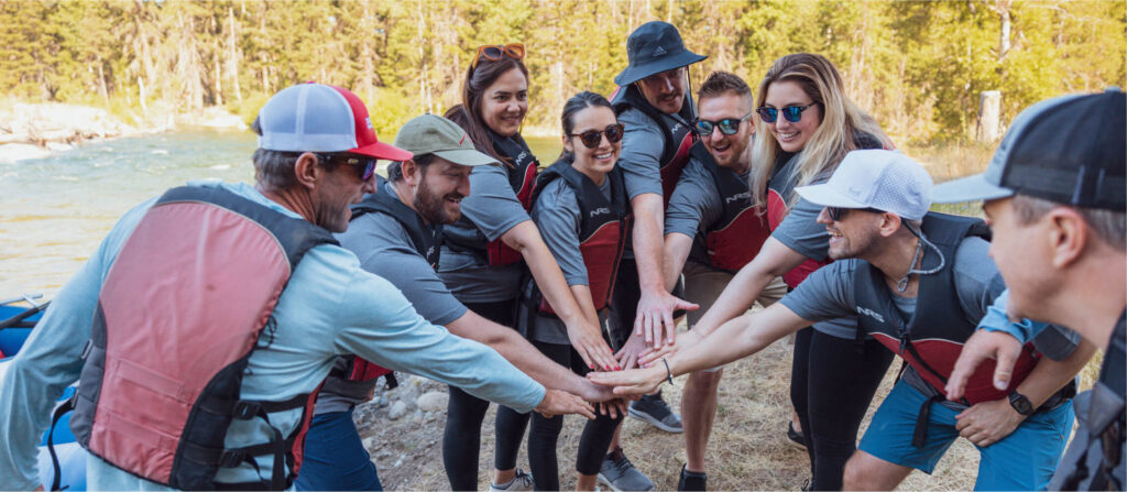 A group of people wearing life jackets stand in a circle with their hands together in the center, outdoors near a river at Suncadia Resort Washington.