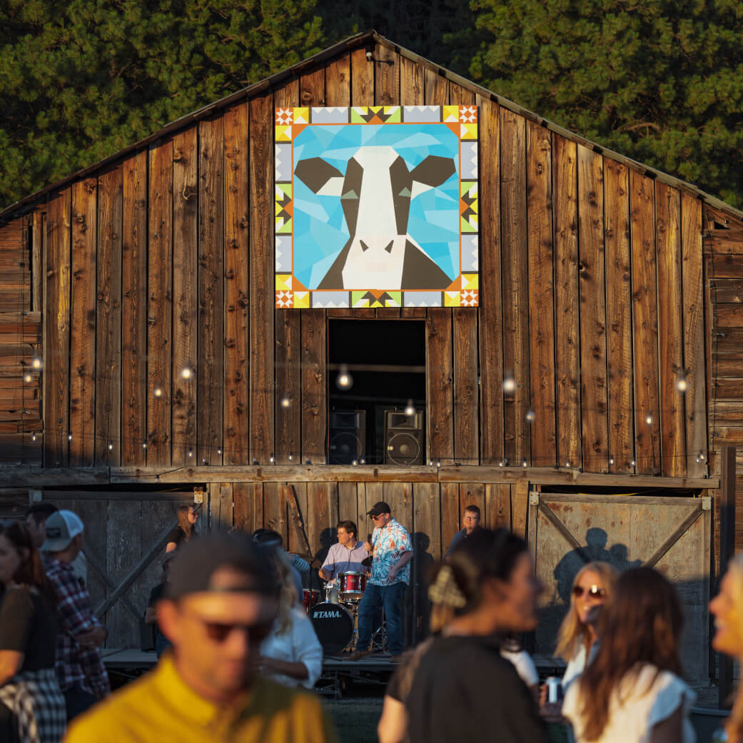 At Suncadia Resort, a wooden barn showcases a striking geometric cow mural. Guests gather outside under the sunny sky, some sipping drinks and enjoying the vibrant atmosphere at this beautiful Suncadia setting.