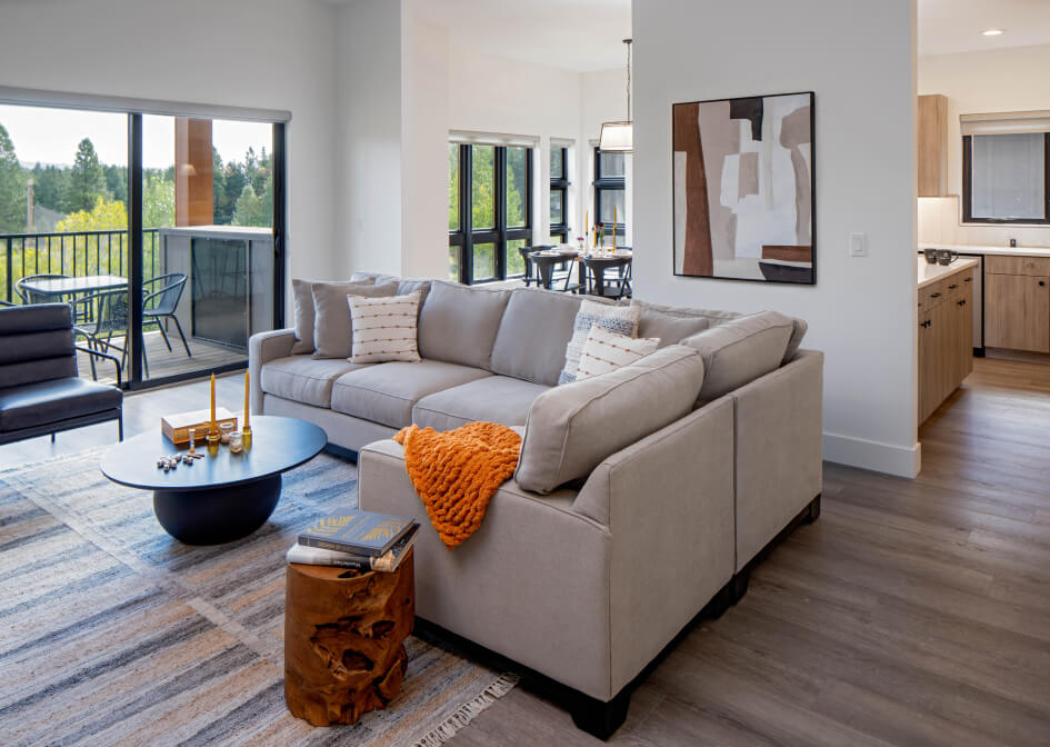 A modern living room in a Suncadia Resort home features a beige sectional sofa, an orange throw blanket, and abstract wall art. Large windows reveal a balcony with lush greenery. The dining area and kitchen are visible in the background, perfect for enjoying after exploring Cle Elum restaurants.