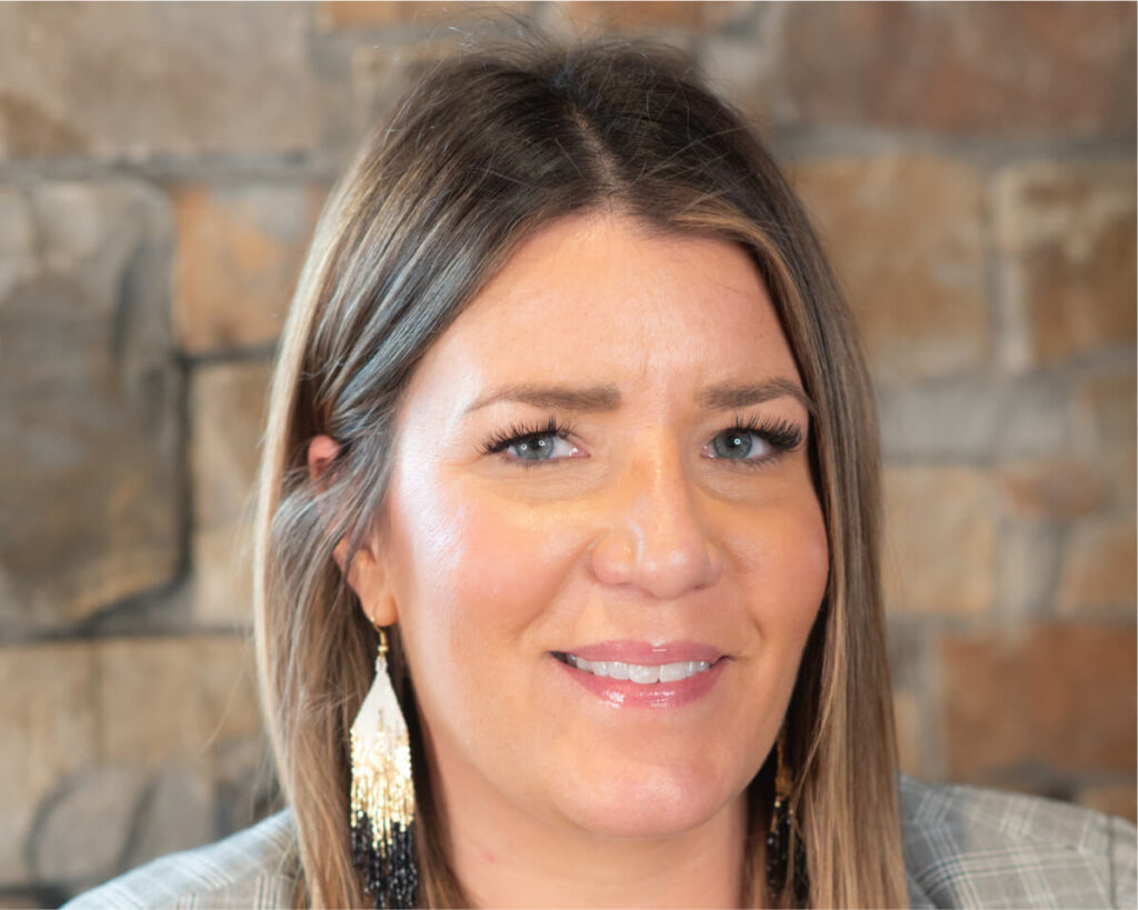 A woman with long brown hair, wearing a plaid shirt and tassel earrings, smiles brightly in front of a stone background, reminiscent of the inviting charm found at Suncadia Resort.