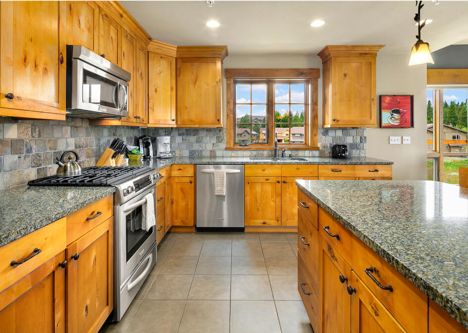 A modern kitchen with wooden cabinets, granite countertops, stainless steel appliances, and a tile floor in a Suncadia home. A window offers natural light and a view of the serene outdoors, capturing the essence of Suncadia Resort Washington living.