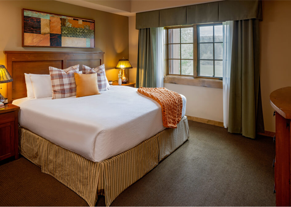 A cozy hotel room at Suncadia Resort features a large bed with multiple pillows, two bedside lamps, a wooden dresser, and a window with green curtains. An orange throw blanket is placed on the bed.