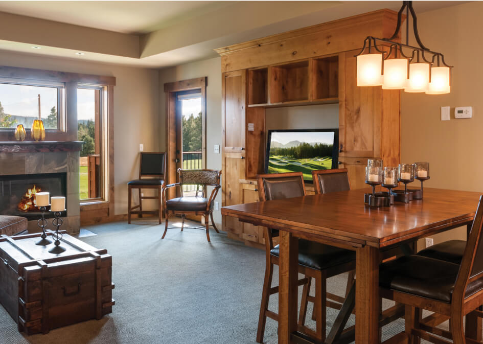 A cozy living room at Suncadia Resort boasts a fireplace, wooden furniture, and a TV. Large windows frame an outdoor view, while in the foreground, a dining table with chairs and a chandelier complete the charming Suncadia homes ambiance.