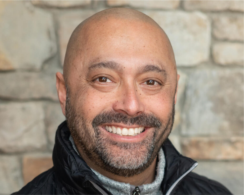 A person with a bald head and beard smiles in front of a stone wall at Suncadia Resort, wearing a black jacket and a light-colored sweater.