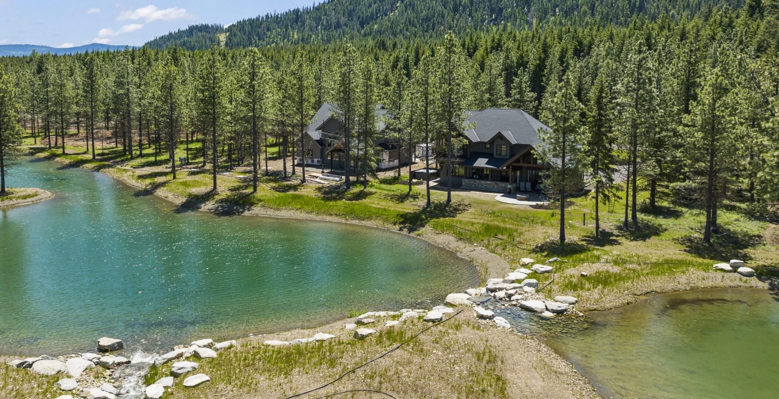 A log cabin house surrounded by tall pine trees sits beside a clear pond with rocky edges in the picturesque Suncadia landscape, just a short drive from charming Cle Elum restaurants.