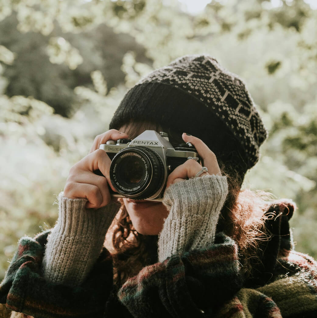 In a leafy, sunlit setting near Suncadia, a person wearing a knitted hat and sweater raises a film camera to their face, capturing the serene beauty of the outdoors.