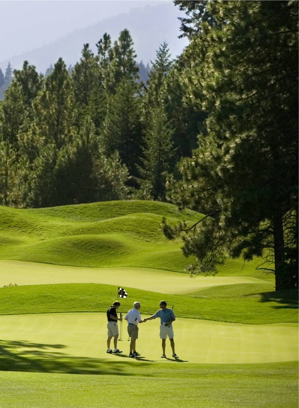 Three people stand on a golf course at Suncadia Resort, two of them shaking hands near a flagstick on a green, surrounded by rolling hills and trees.