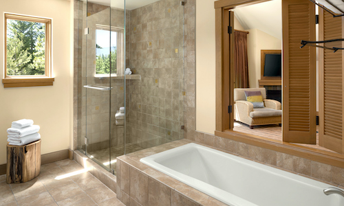 A bathroom at Suncadia Resort features a tiled bathtub and glass shower, offering a view into the living area with a chair and TV. A stack of towels rests on a wooden stool near the shower, adding comfort to your stay after enjoying nearby Cle Elum restaurants.