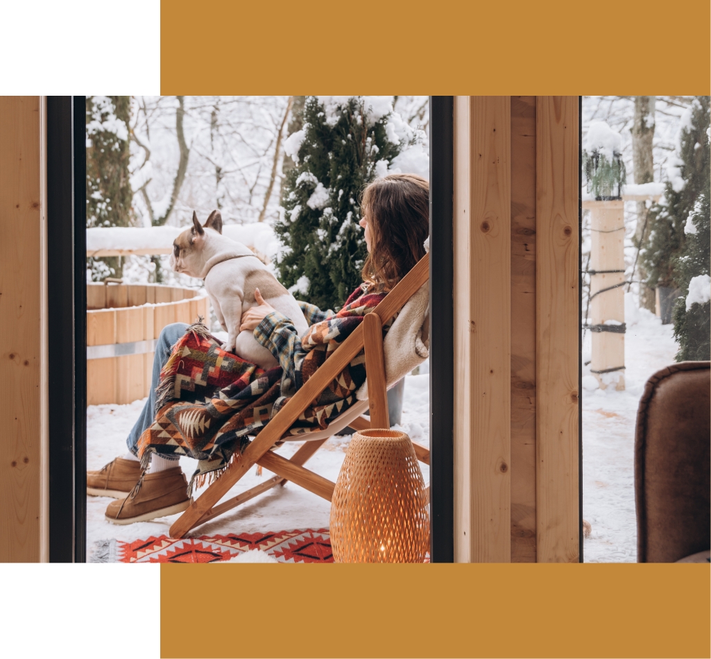 Wrapped in a cozy blanket, a person sits in a deck chair on a snowy patio at Suncadia Resort, gently holding their dog. Surrounded by serene trees, they enjoy the tranquil scenery that makes this place feel like home.