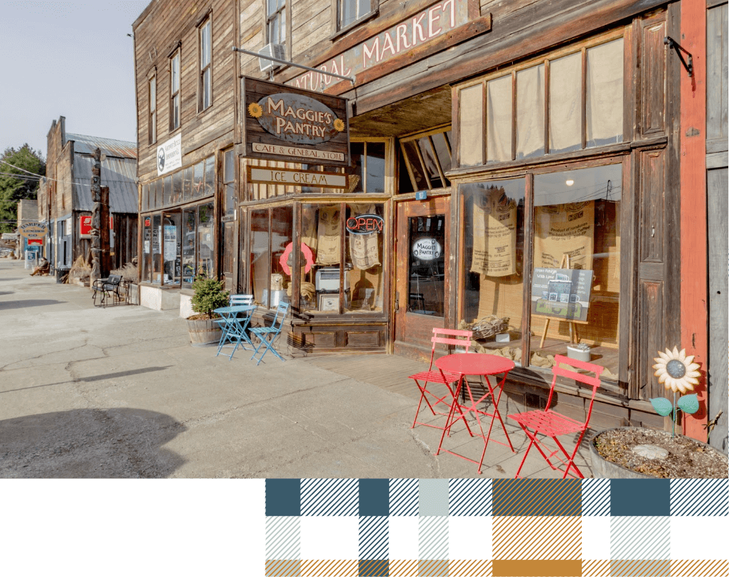 A quaint street with wooden buildings features a store called Maggie's Pantry. Red and blue chairs sit outside, surrounding a small round table under a sign that says "open." This charming spot, reminiscent of the Cle Elum restaurants near Suncadia homes, invites passerby to relax and enjoy.