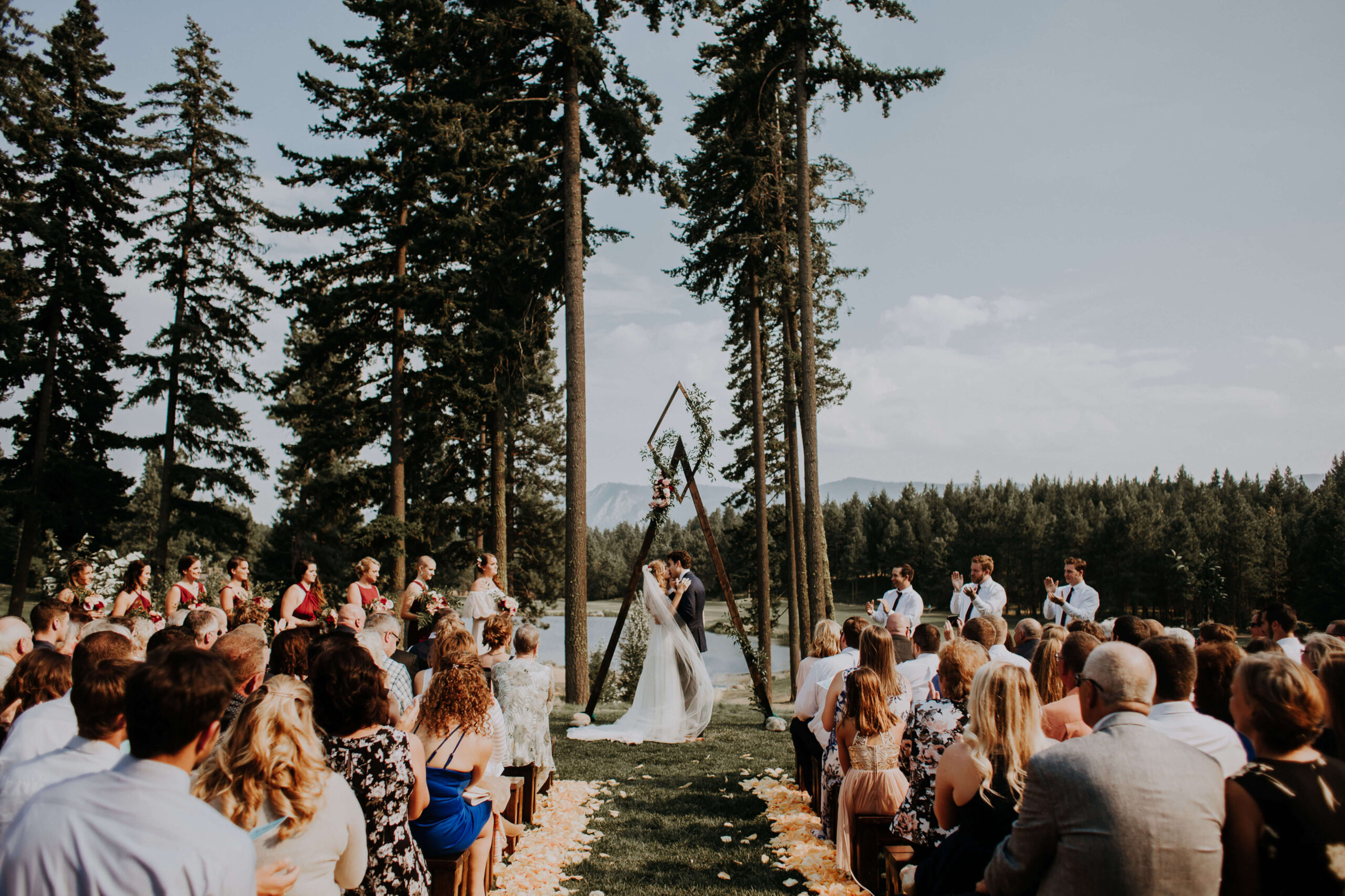 An outdoor wedding ceremony unfolds under a geometric arch, with the couple surrounded by guests seated on either side. Tall trees and a breathtaking mountainous landscape of Suncadia Resort in Washington provide a stunning backdrop.