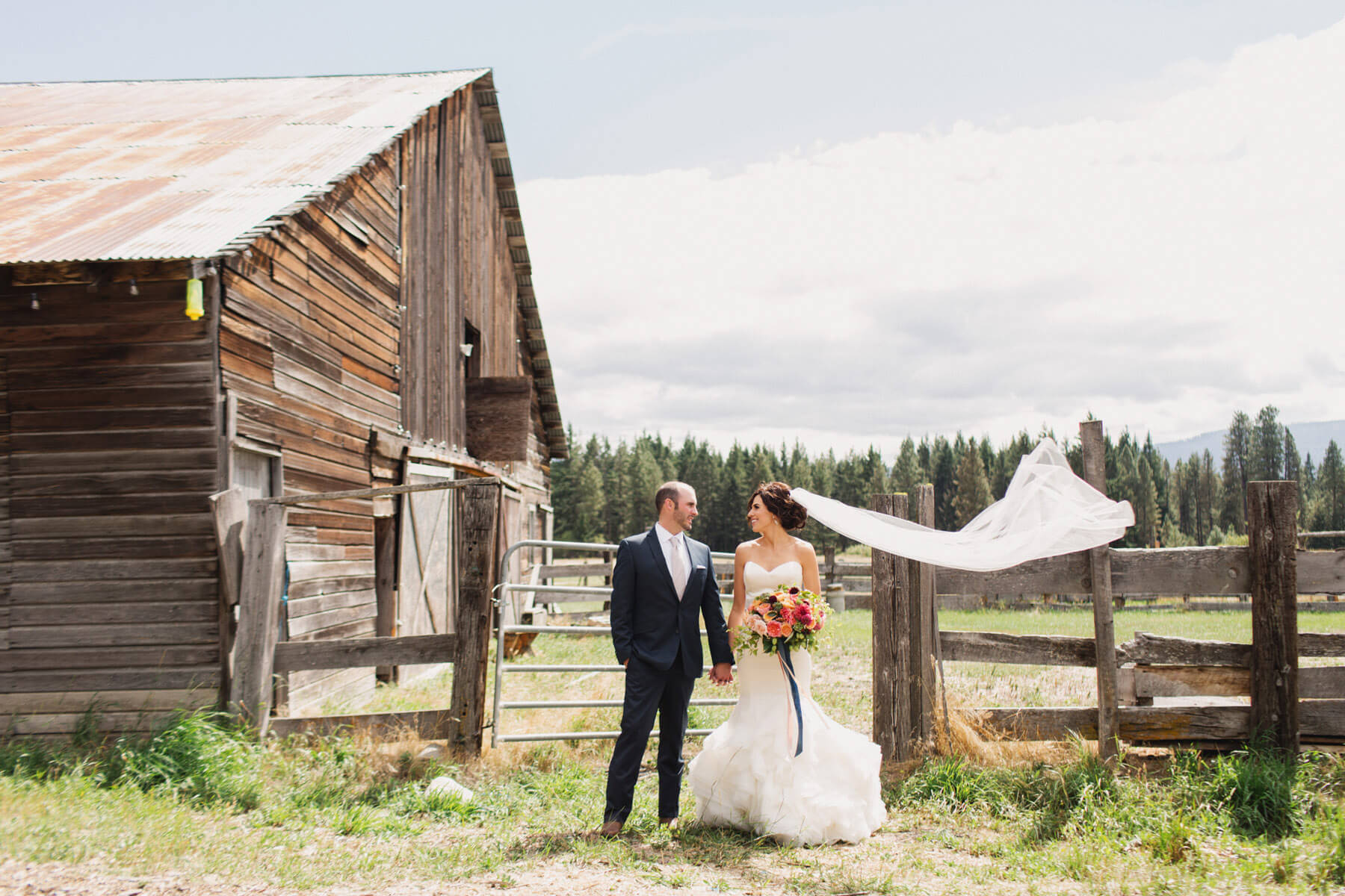 A bride and groom stand holding hands outside a rustic wooden barn at Suncadia Resort. The bride’s veil flows in the wind as she holds a bouquet, embodying the enchanting charm of this Washington venue.