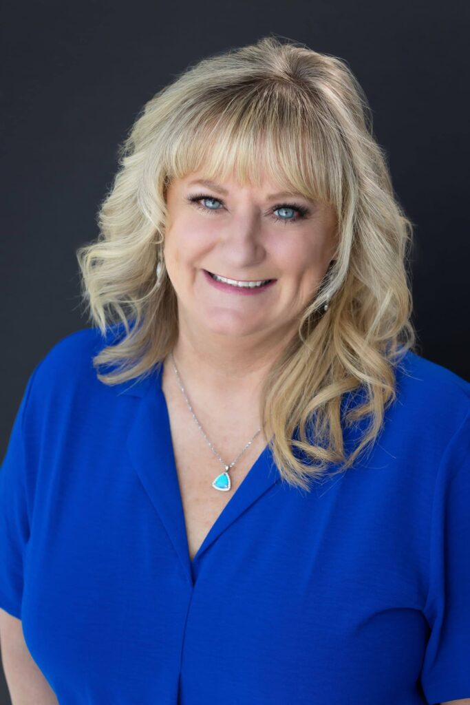 A person with long blonde hair wearing a blue shirt smiles, capturing the warmth and relaxation reminiscent of Suncadia Resort's inviting atmosphere, standing against a dark background.