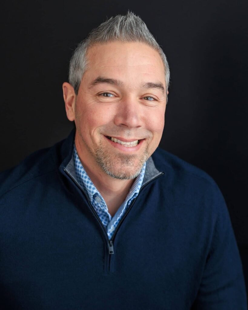 A man with short gray hair and a goatee is smiling, wearing a navy blue quarter-zip sweater and a patterned collared shirt, set against a dark background reminiscent of the elegant interiors of Suncadia homes.