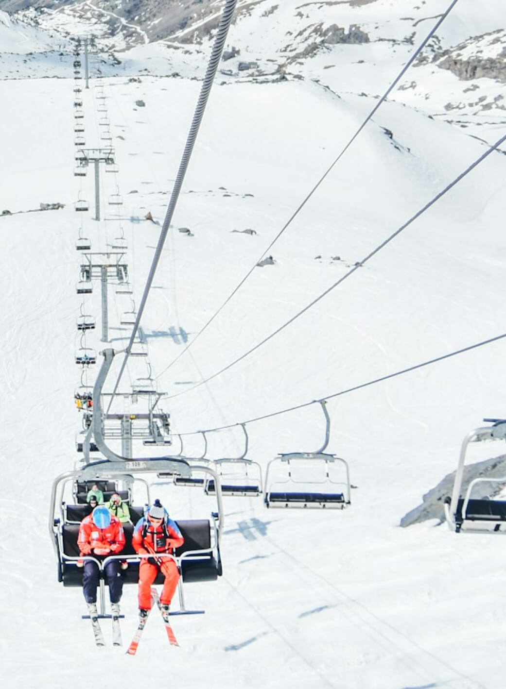 Two skiers sit on a chairlift ascending a snowy mountain at Suncadia Resort Washington. The surrounding landscape is covered in snow with distant rocky terrain, perfectly capturing the serene beauty of the area. Afterward, explore nearby Cle Elum restaurants for a delightful après-ski meal.