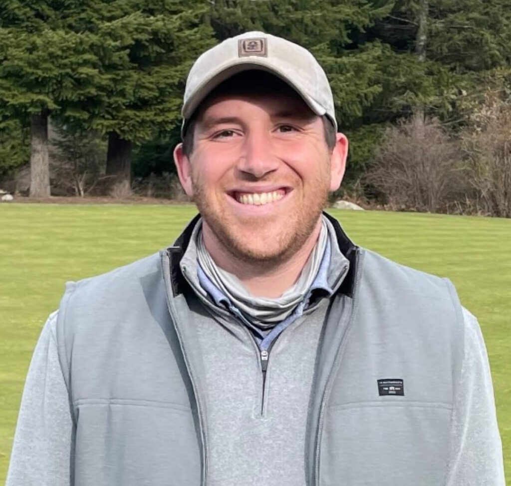 A person in a gray vest and cap smiles while standing outdoors on a grassy area with trees in the background.