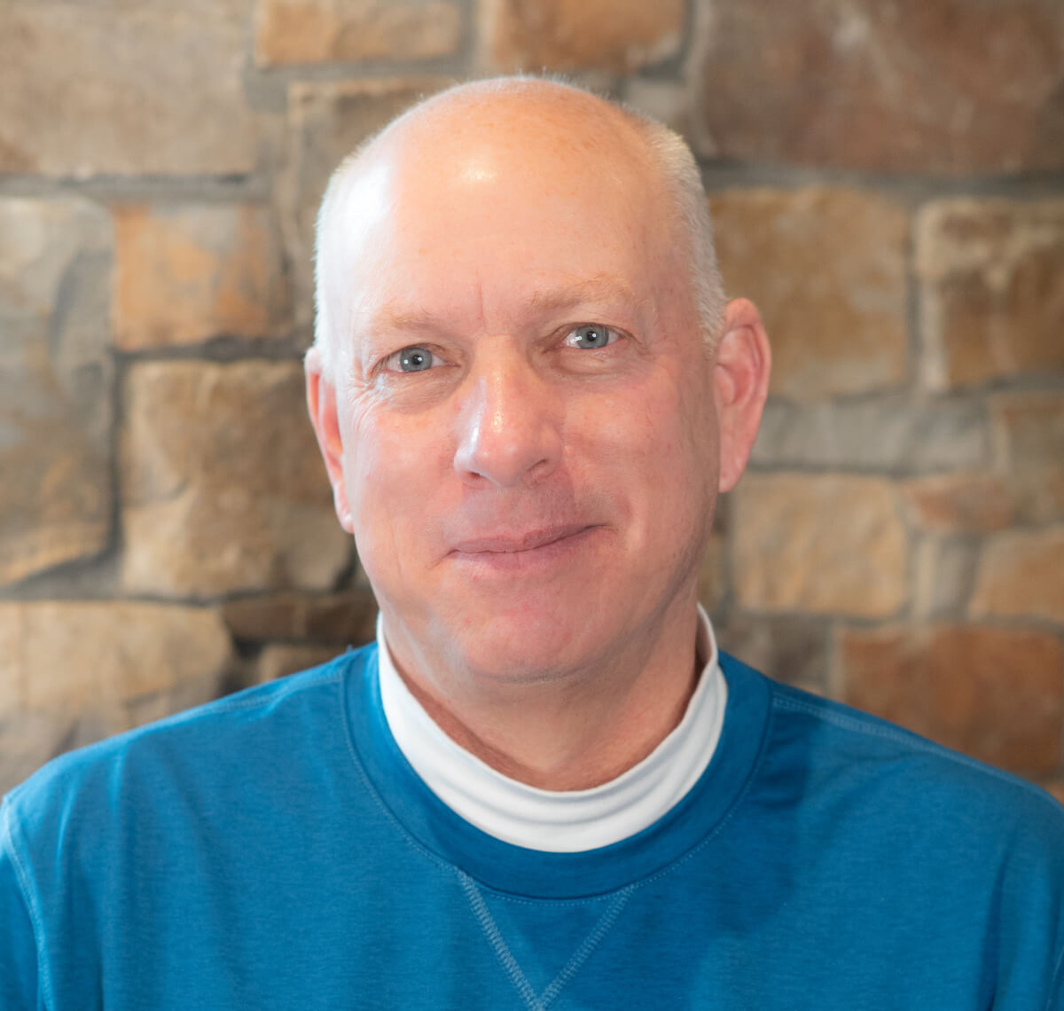 A bald man in a blue shirt smiles in front of a stone wall backdrop.