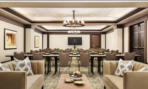 A conference room with rows of chairs and tables facing a screen. The room is decorated with artwork on the walls, a chandelier, and cushioned couches with patterned pillows in the foreground.