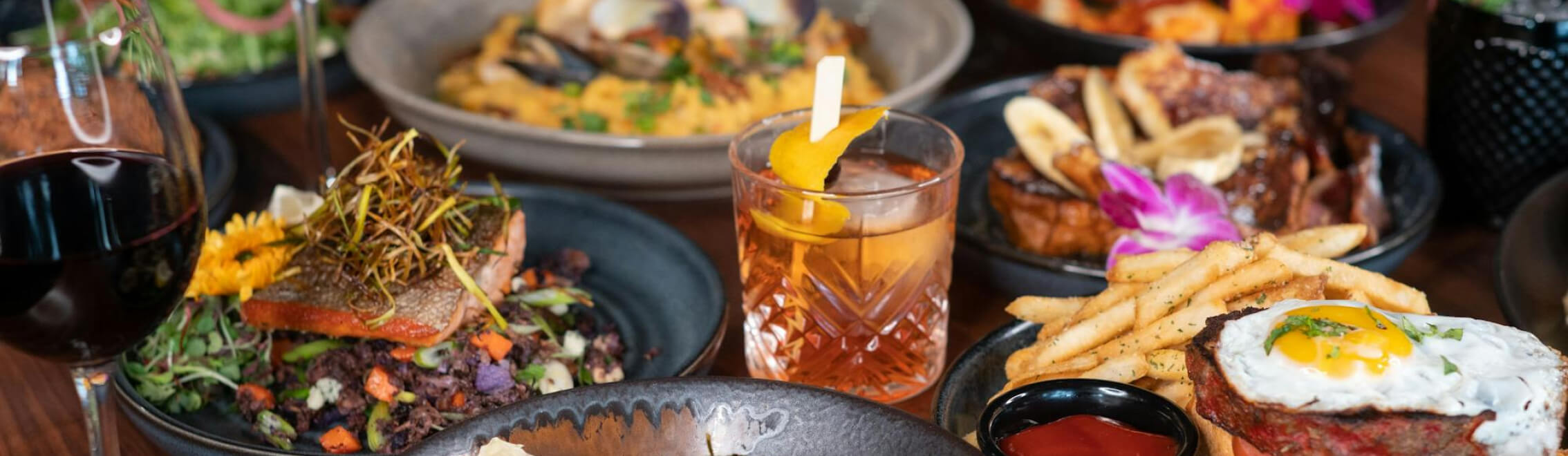 A table with assorted dishes and drinks, including seafood, steak with fries, a cocktail, and a glass of red wine.