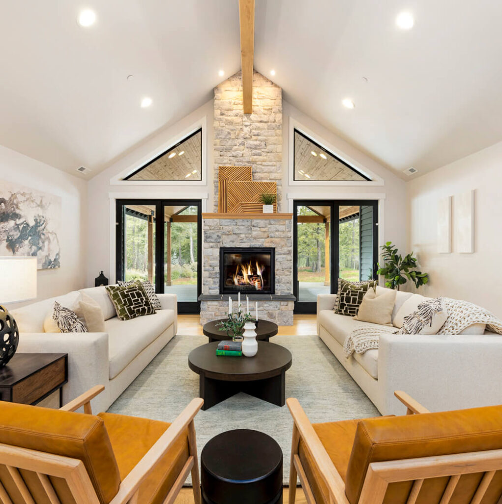 Stylish living room with vaulted ceiling, stone fireplace, two cream sofas, and two wooden chairs. Large windows offer a view of trees.