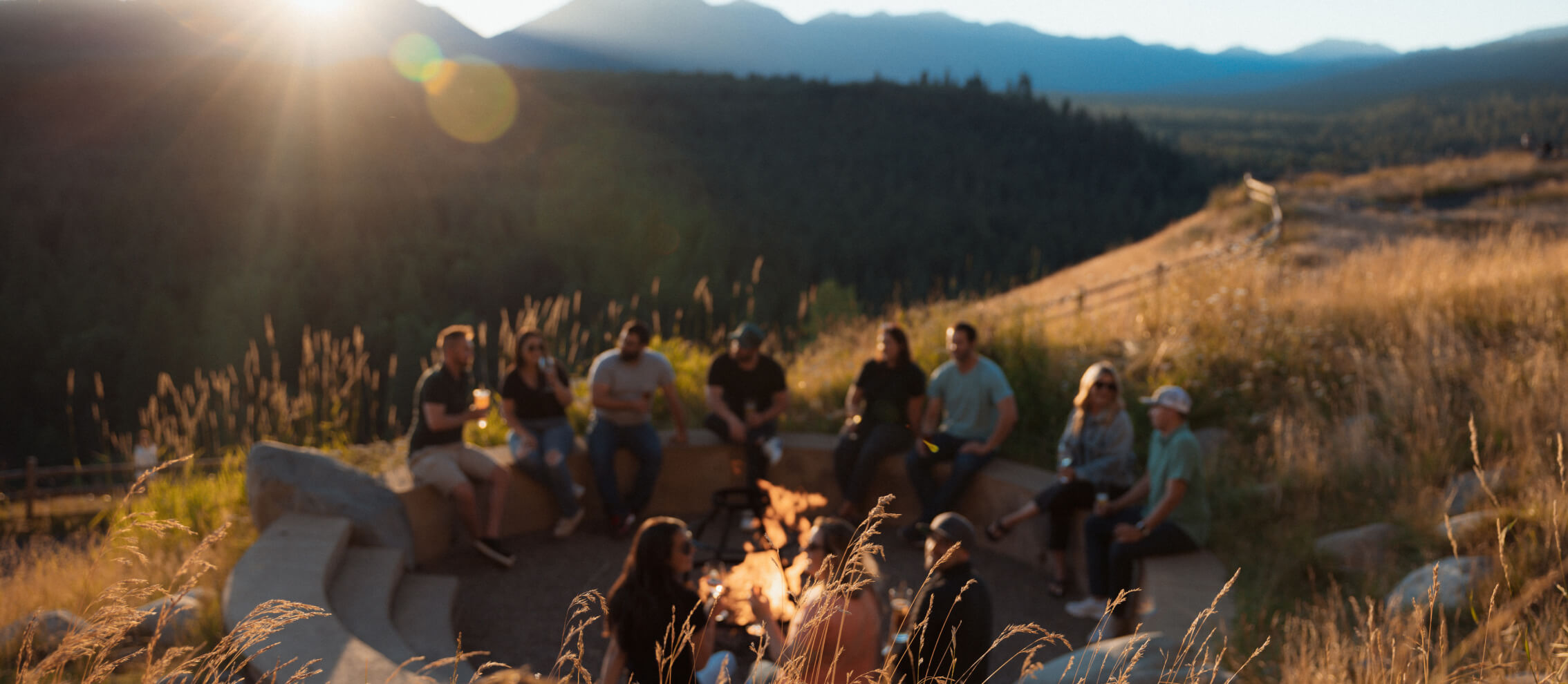 A group of people sitting around a campfire in an open field with mountains in the background under a setting sun.