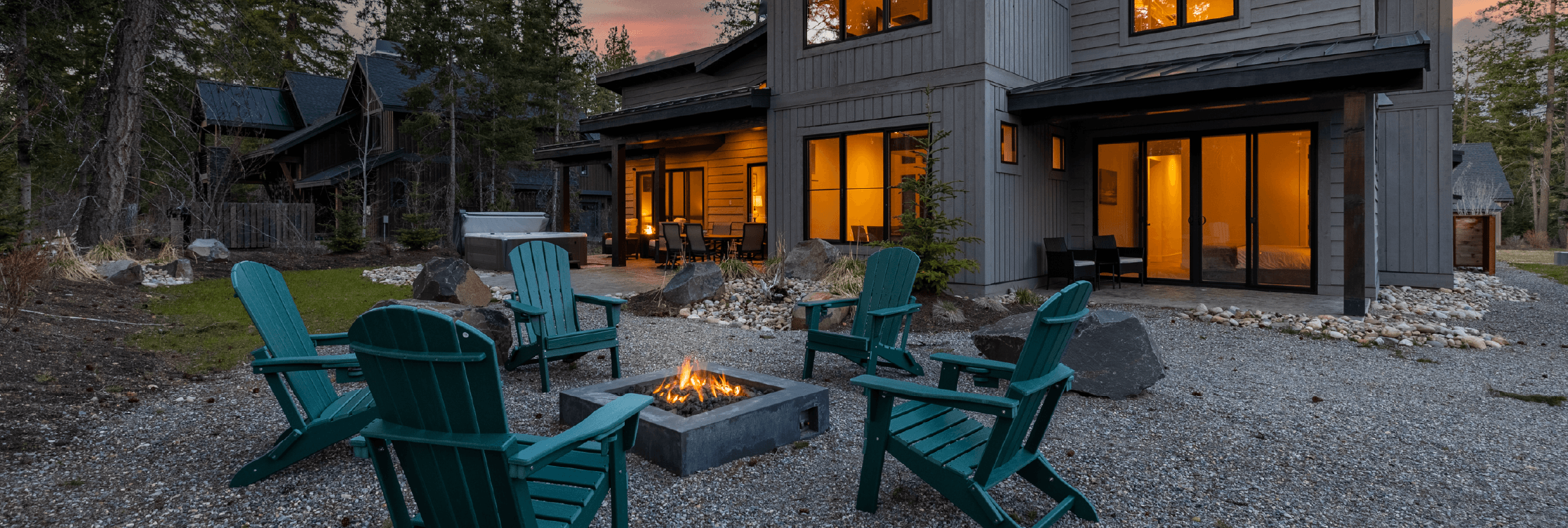 A modern house with large windows sits behind a gravel patio with six teal Adirondack chairs arranged around a square fire pit at sunset, surrounded by trees and rocks.