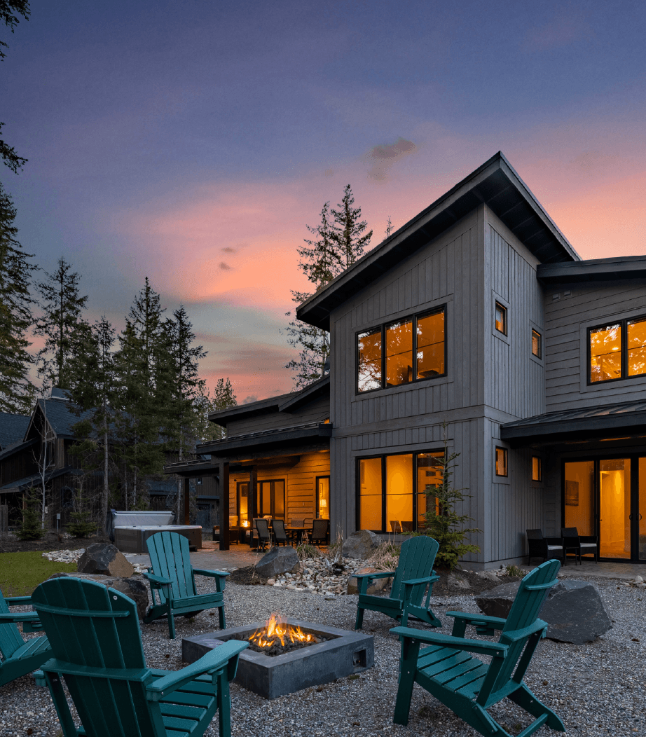 Modern two-story house at dusk with warm lights on, surrounded by trees. Green Adirondack chairs circle a lit fire pit in the gravel yard, creating a cozy outdoor space under a colorful sunset sky.