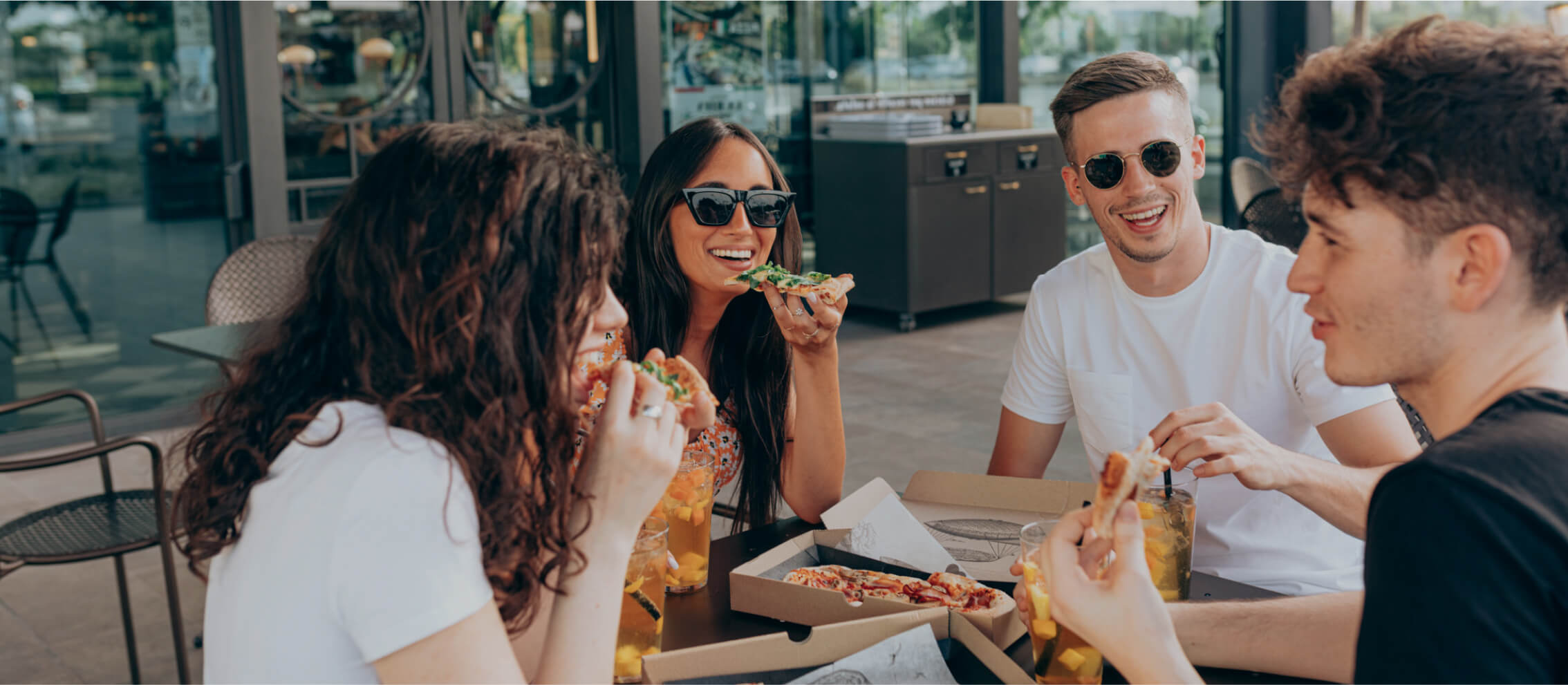 Four people sitting outdoors, wearing casual attire and sunglasses, eating pizza and smiling.