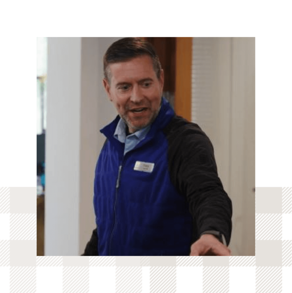 Man in a blue vest speaking and gesturing, standing indoors.