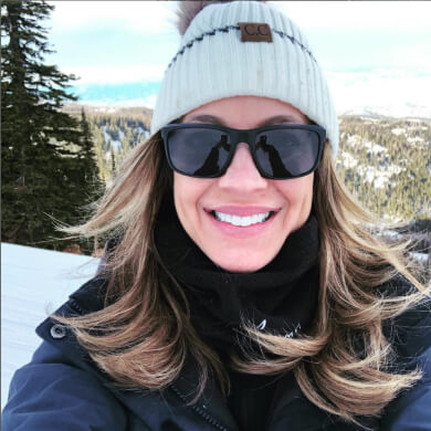 Person wearing a white beanie and sunglasses, smiling outdoors in a snowy, mountainous area with pine trees in the background.