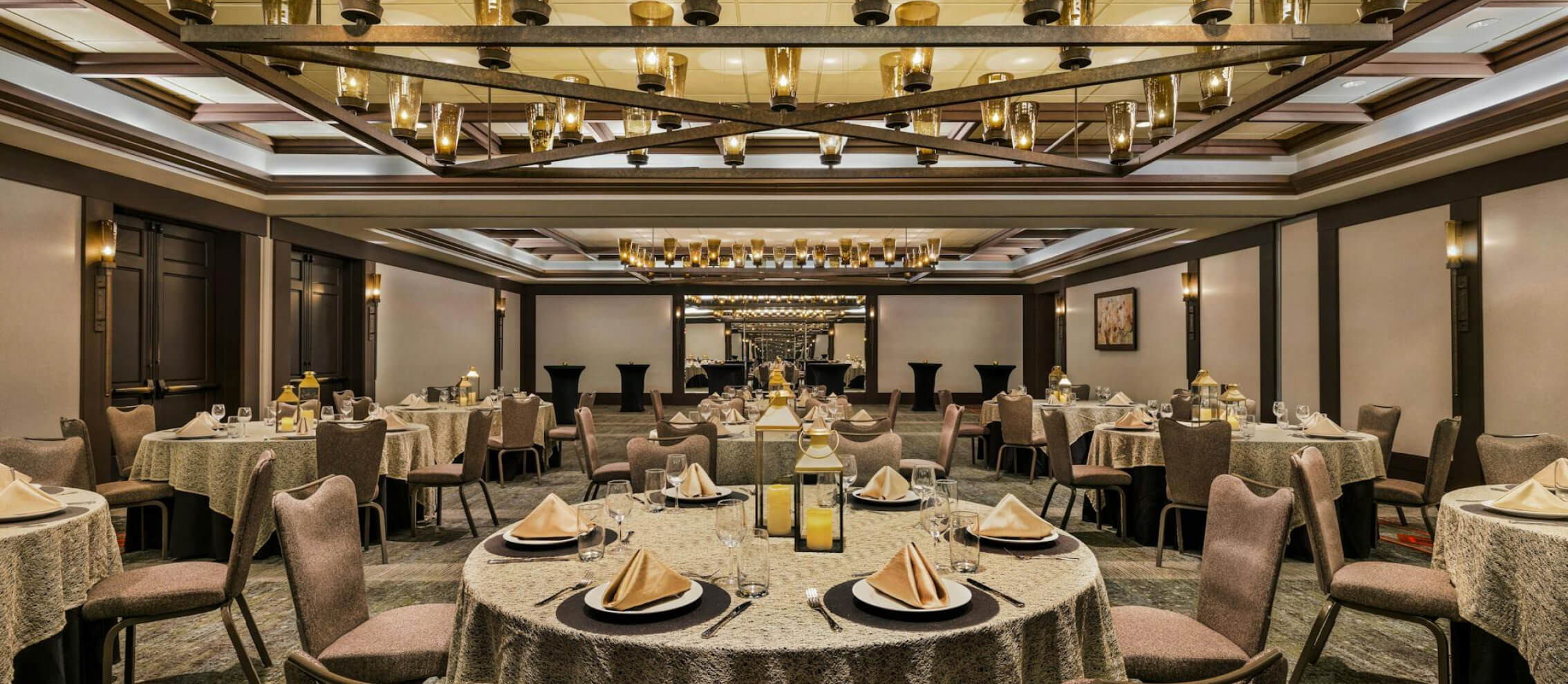 Elegant banquet hall set with round tables, beige tablecloths, folded napkins, and glassware. Overhead lighting features modern chandeliers and a large mirror at the back wall.