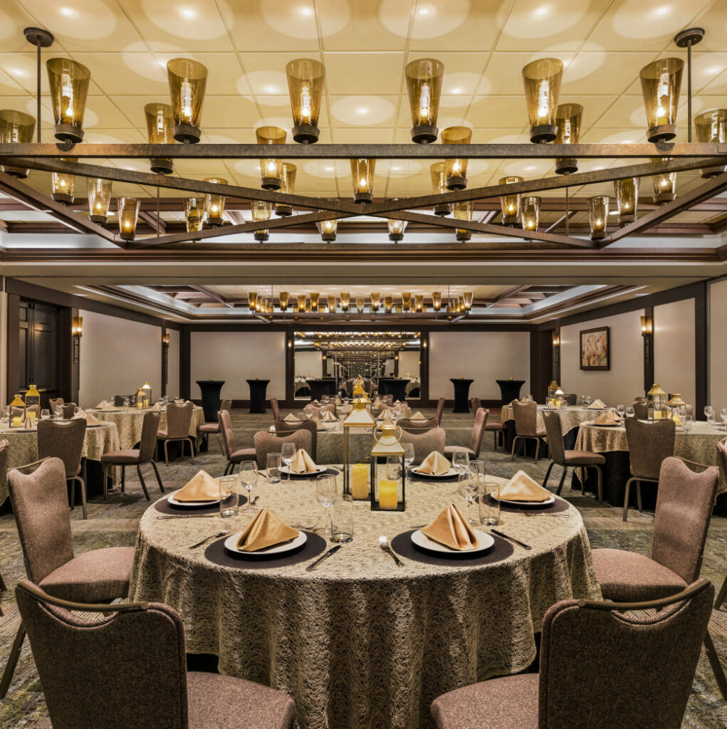 Elegant banquet hall with round tables set with tableware and napkins. Chandeliers hang from the ceiling, and mirrors adorn the walls.