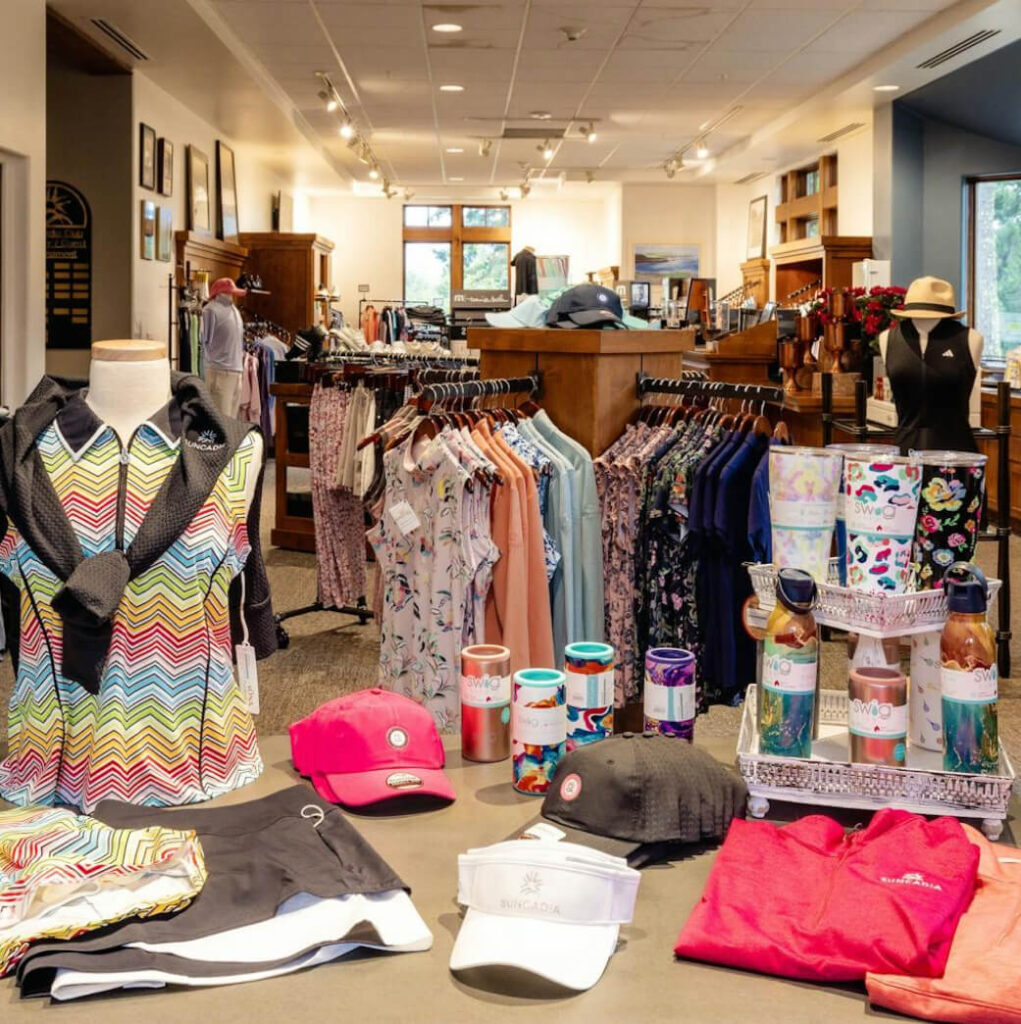 A clothing store display with colorful apparel on hangers, a table with hats, drinkware, and folded garments. Bright and organized interior with natural light from large windows.