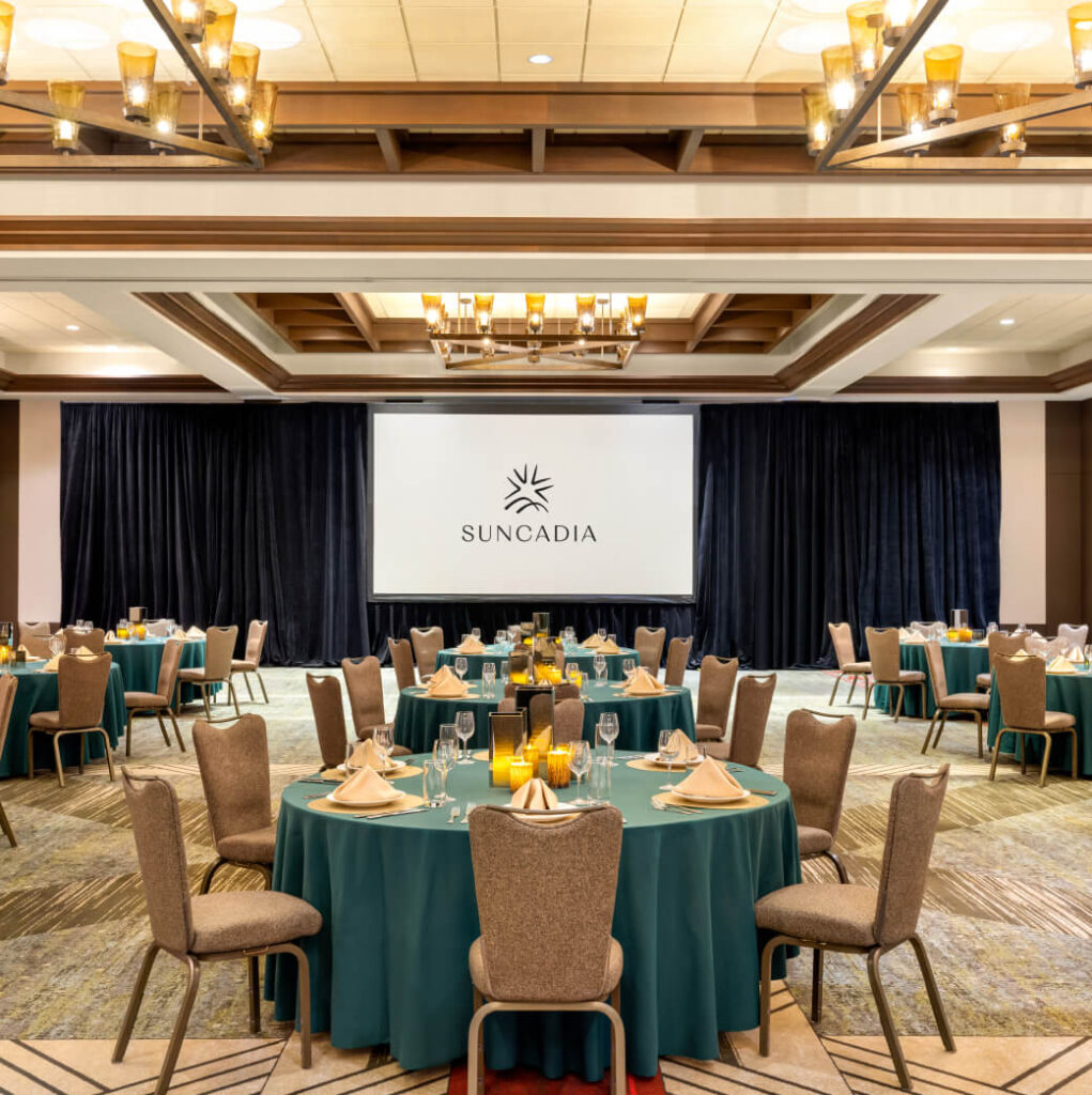 A conference room set up with round tables covered in teal tablecloths. Each table has place settings and decorative candles. A large screen at the front displays the Suncadia logo.