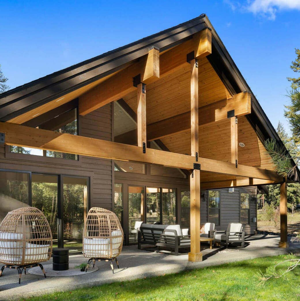 A modern wood and glass house features a sloped roof, large windows, and a spacious outdoor patio with wicker chairs and a sofa set, surrounded by trees.