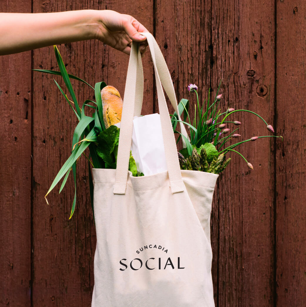 A hand holding a tote bag with the words "Suncadia Social" printed on it. The bag contains a baguette, leafy greens, and purple flowers, in front of a wooden wall.