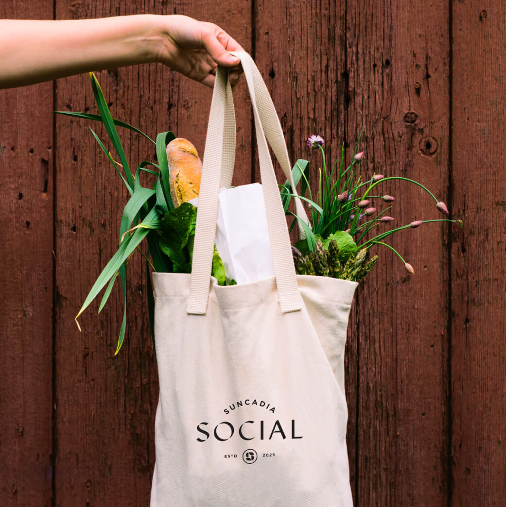 A hand holds a tote bag with the text "Suncadia Social." The bag contains a baguette, green vegetables, and potted plants against a wooden background.