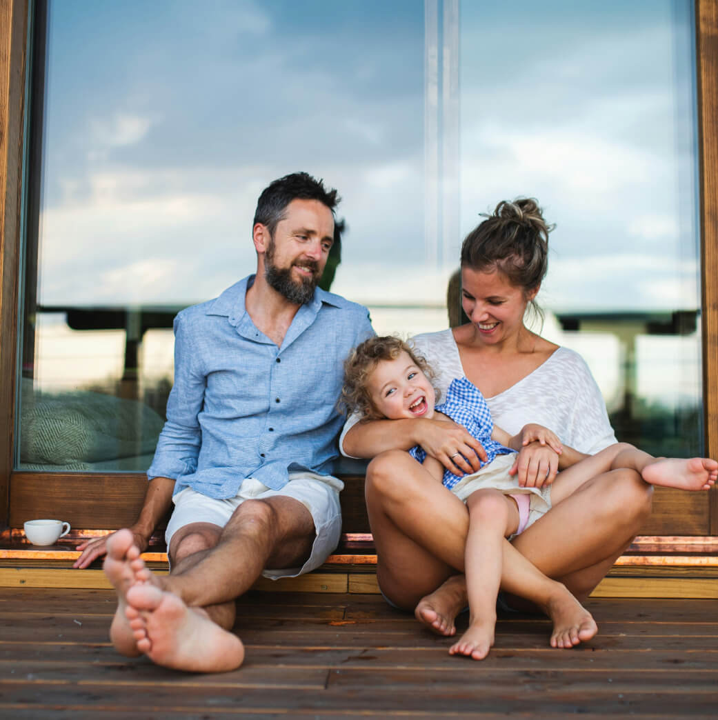 A family of three sits on a wooden porch; the parents smile at their child, who is seated on the mother's lap. A coffee cup is on the floor beside them.