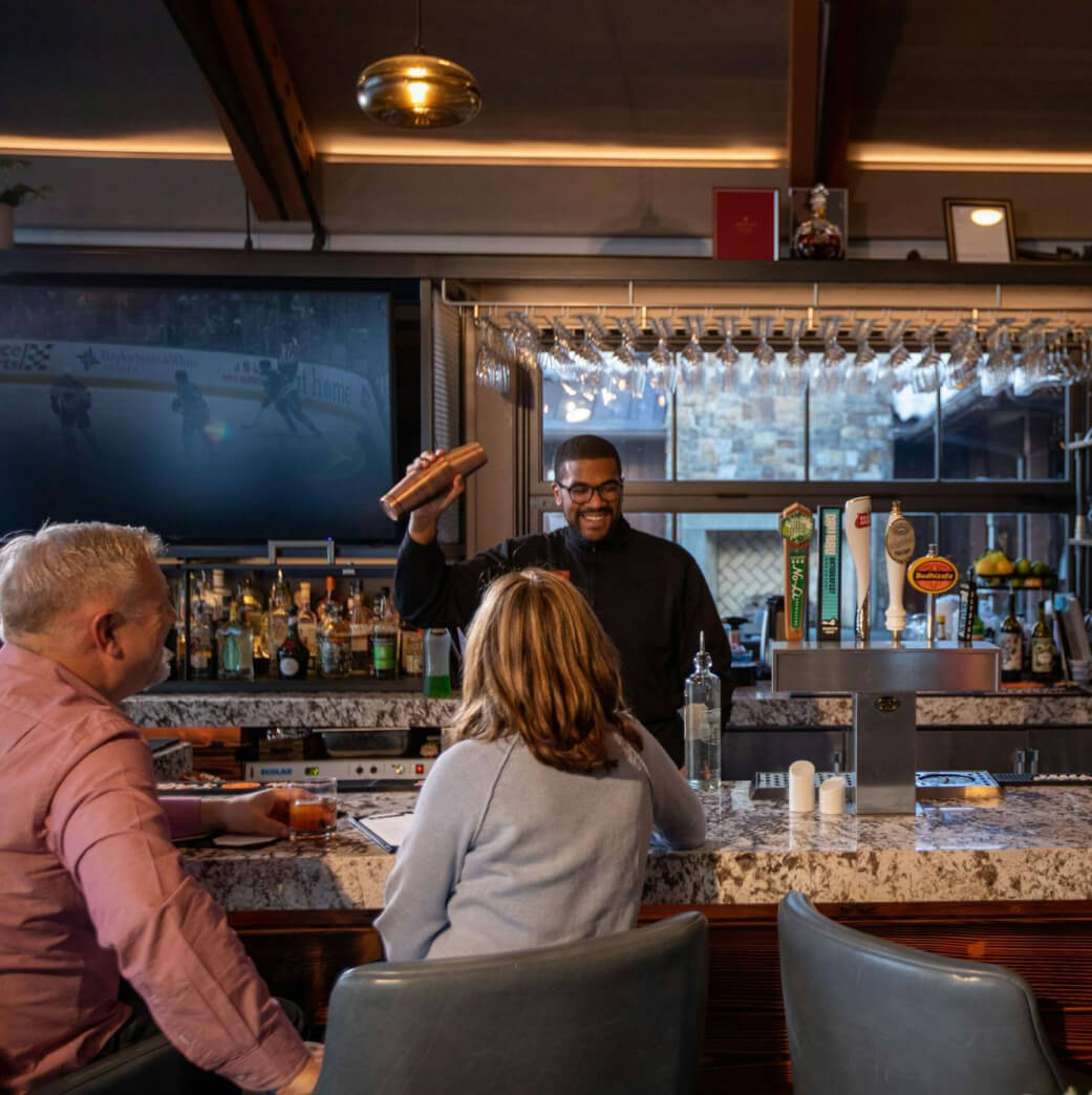 A bartender shakes a mixer behind a bar with various taps, while two people sit on stools, conversing. A TV in the background displays a sports event.