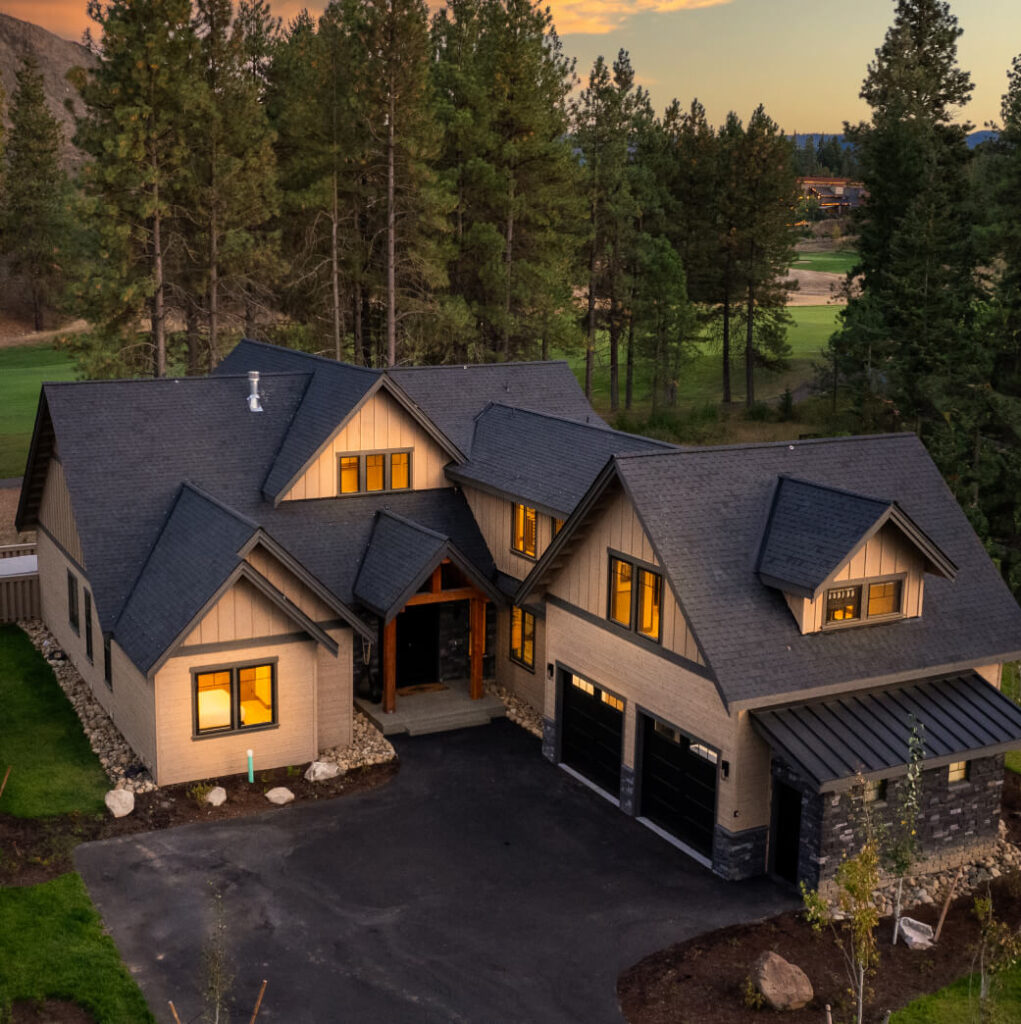 A large modern home with multiple gabled roofs, surrounded by trees and a paved driveway in front. The sky is clear with a hint of sunset in the background.