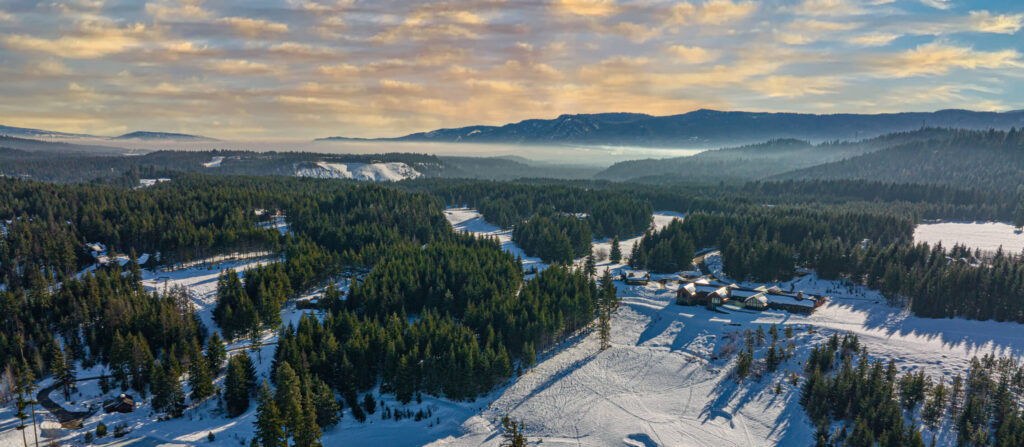 Aerial view of a snowy forest and hills with scattered buildings under a cloudy sky at sunset.