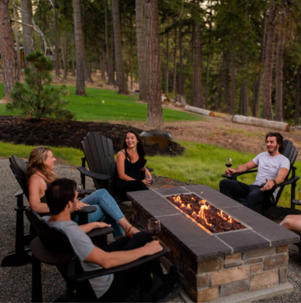 Four people sitting around an outdoor fire pit, holding drinks, surrounded by trees.