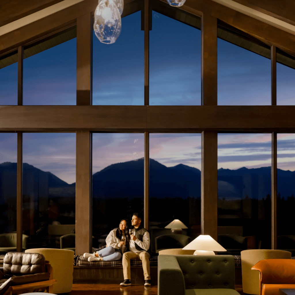 A couple sits by large glass windows in a wooden cabin, looking out at a mountain range during sunset. Cozy furniture surrounds them, and a lamp is lit nearby.