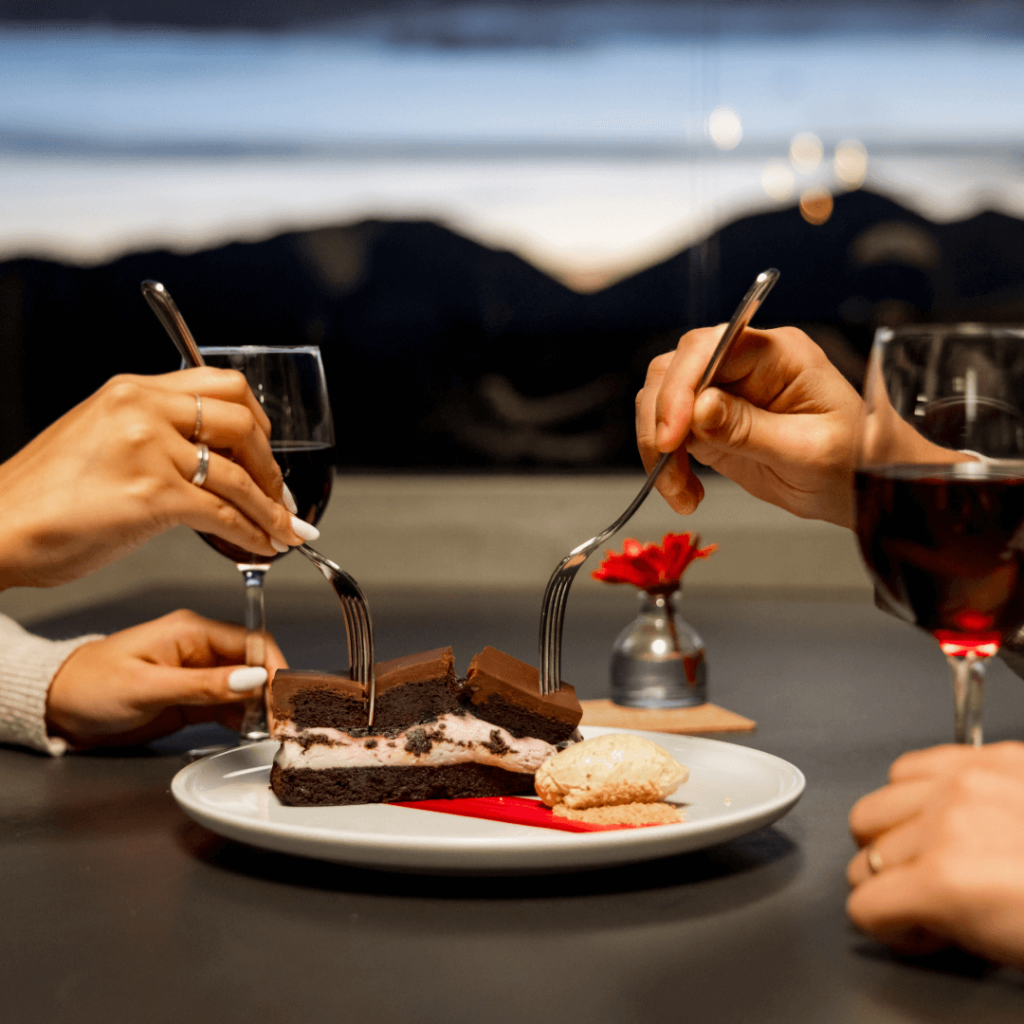 Two people enjoy chocolate cake with forks, next to glasses of red wine on a table. A small red flower in a vase is in the background, and a twilight landscape is visible outside.