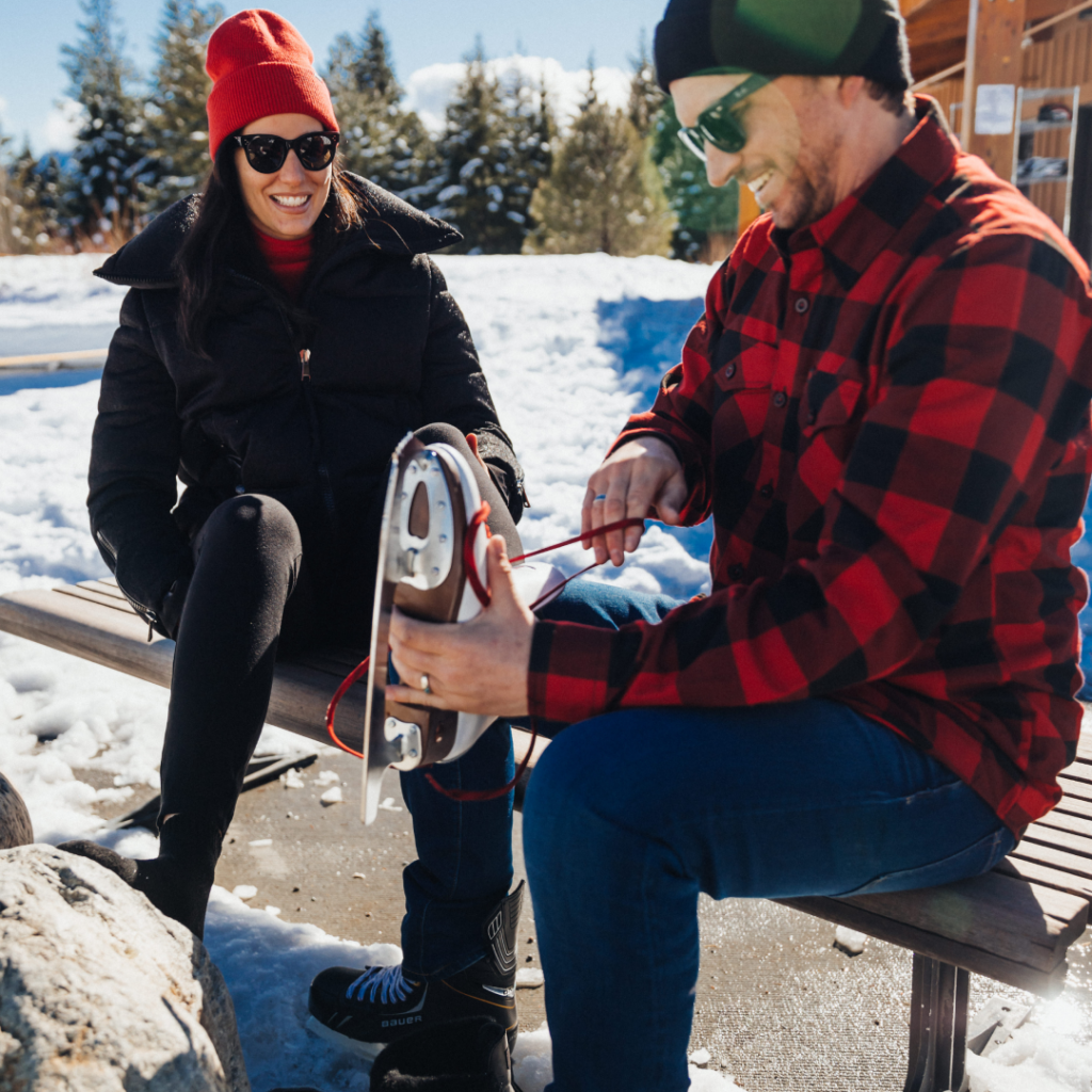 Two people sitting on a bench in a snowy setting, with one person tying ice skates on the other's foot. Both are dressed in winter clothing.