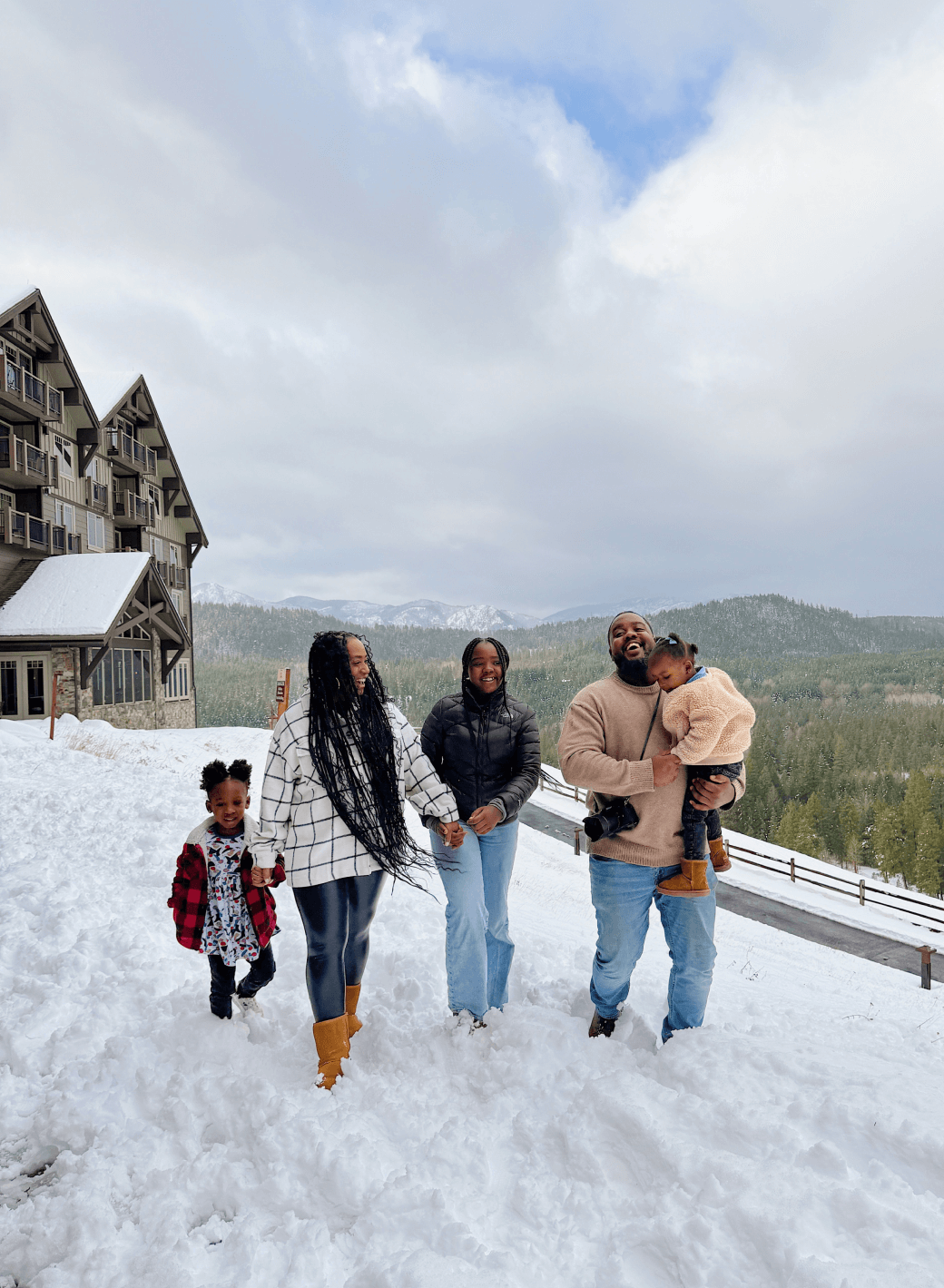 A family of four walks in the snow near a wooden lodge, surrounded by a snowy landscape and mountains in the background.