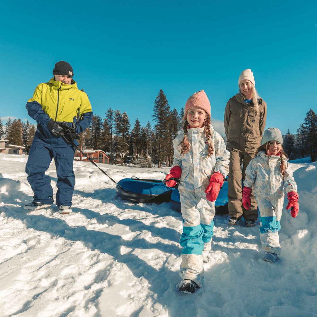 A family of four enjoys a snowy day. Two adults stand while two children walk in the snow, dressed in winter gear. A sled is on the ground. A clear blue sky and trees are in the background.