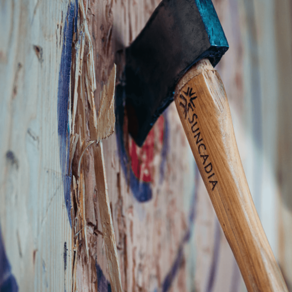 A Suncadia-branded axe is lodged in a wooden target, surrounded by chipped wood and paint marks.